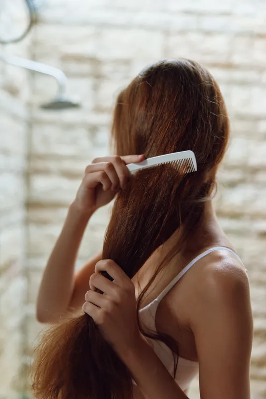 Young woman combing long wet hair in the shower, natural light highlighting her skin and hair strands, bathroom background with blurred tiles.