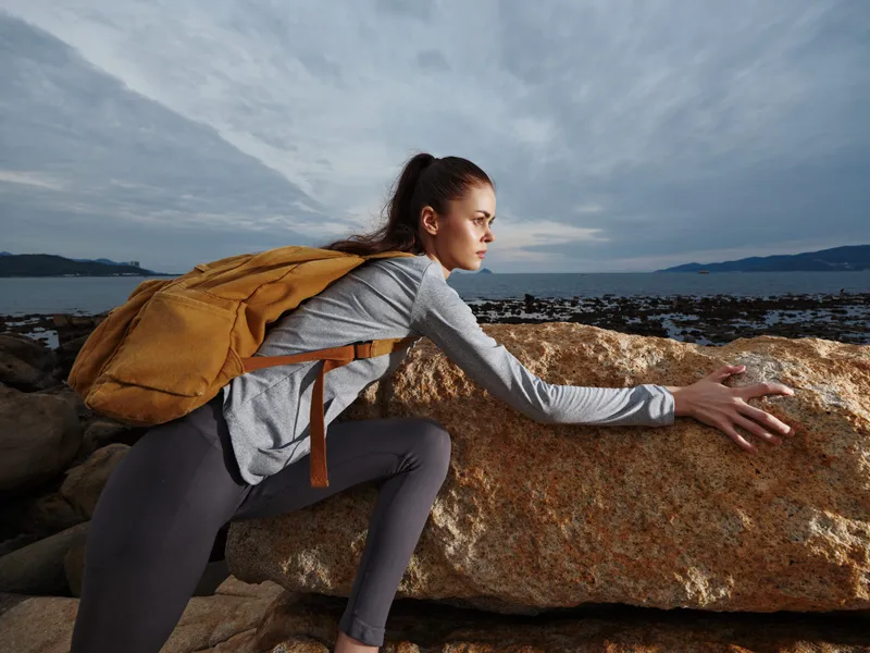 Young woman climbing a rock at the beach, showcasing determination and outdoor adventure spirit with a backpack in a scenic coastal landscape