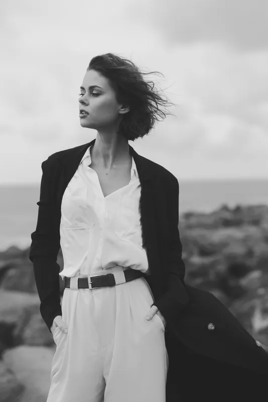 Wind swept fashion on a coastal scene, monochrome styling of a woman in blazer and white shirt with waist trousers, exuding confidence, elegance, and a timeless dramatic mood by the sea