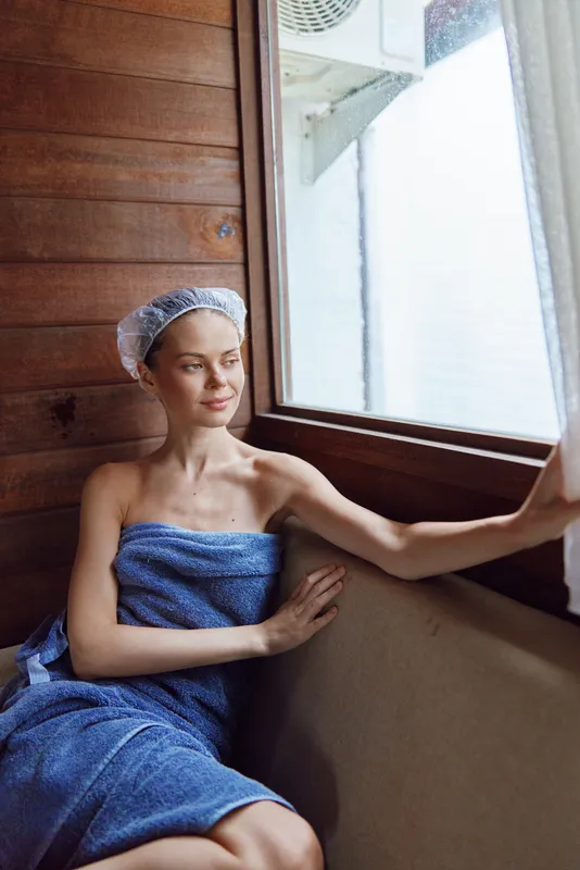 relaxed woman in towel and shower cap sitting by wooden window inside sauna, enjoying peaceful moment with natural light and warm interior tones