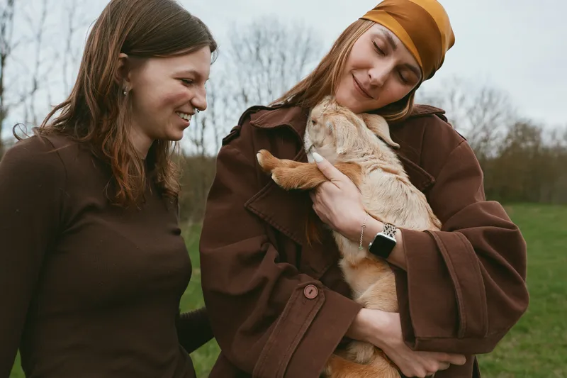 Two women smile outdoors as one cradles a young goat kid in a brown coat, conveying gentle companionship, rural lifestyle, and compassionate animal bond in a sunny field.