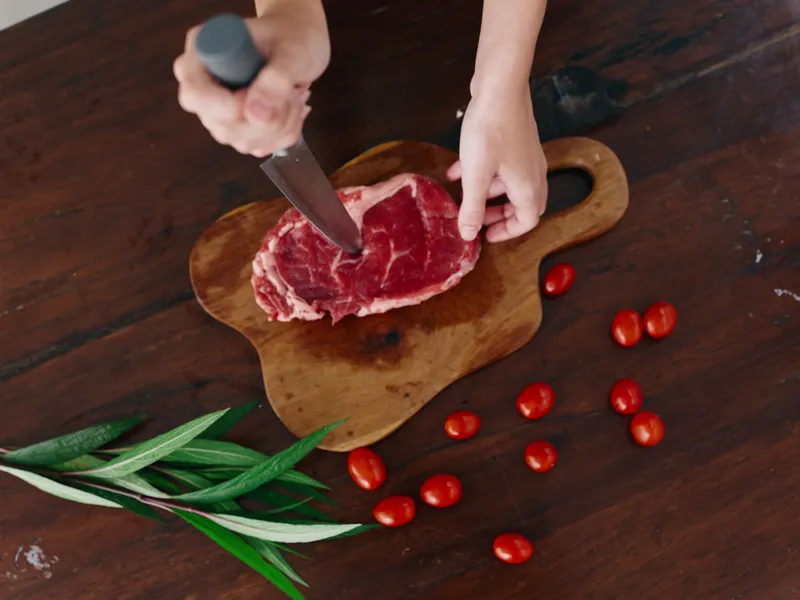 Женщина с ножом в руке нарезает свежее стейковое мясо для жарки на кухне, на столе соль, перец и другие специи, красные помидоры черри и зелень, приготовление ужина