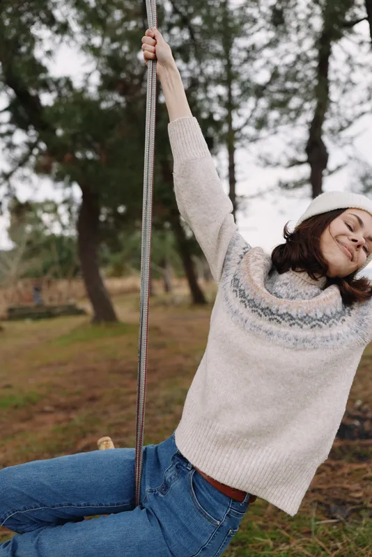 A girl on a rope swing in a park, wearing a cozy sweater and jeans, smiles as she swings through a breezy autumn day in a lush outdoor setting.