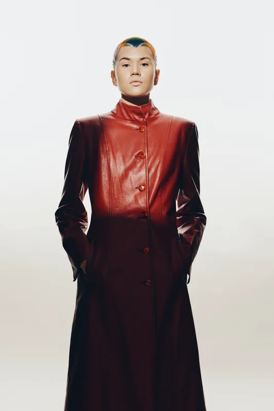 Woman in long dark red leather coat standing with hands in pockets, short hair, neutral expression, minimalist background, fashion style, modern look.