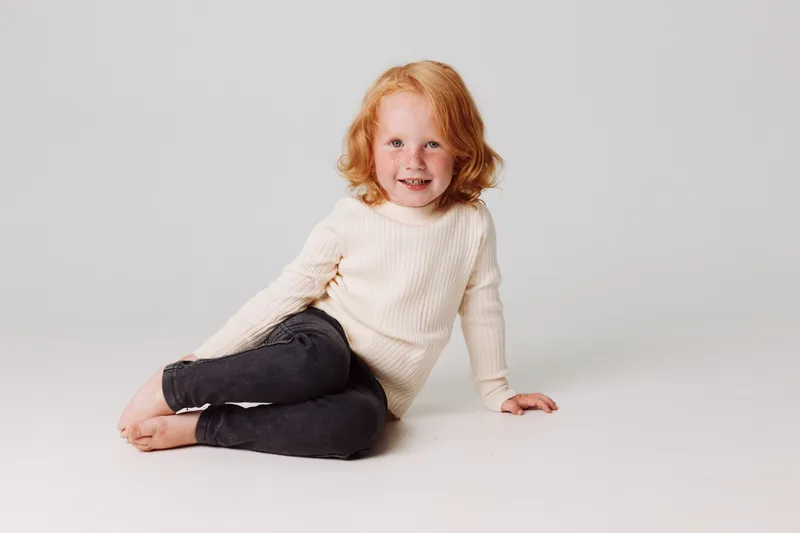 Юная девушка в белой рубашке и черных леггинсах сидит на полу в удобной позе дома