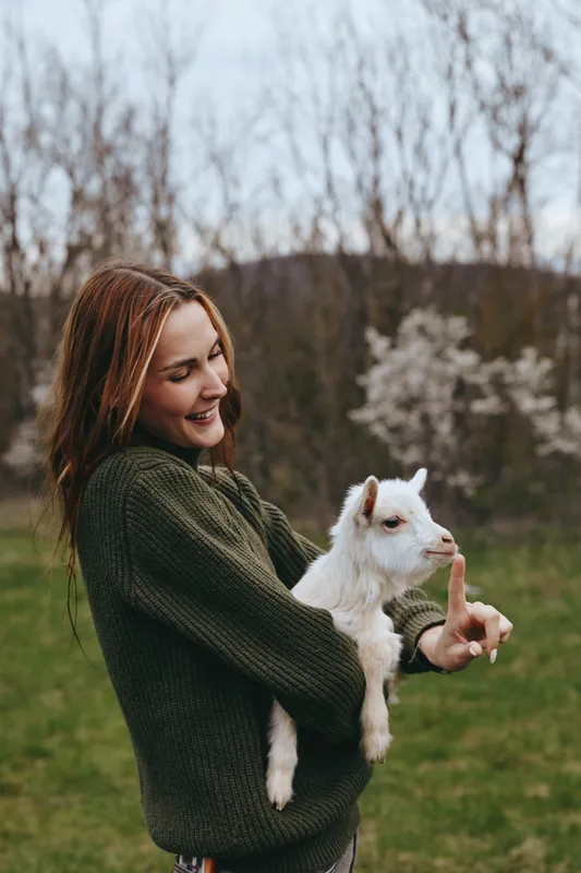 Lifestyle outdoors with a smiling woman holding a white goat kid in a sunny field, casual sweater and gentle interaction, candid moment, natural colors, lush grass and trees in the background