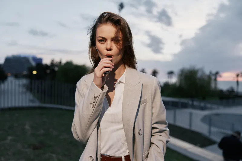 Woman in beige jacket and white blouse expressing thoughtful emotion outdoors at dusk, urban background with soft natural lighting and muted colors