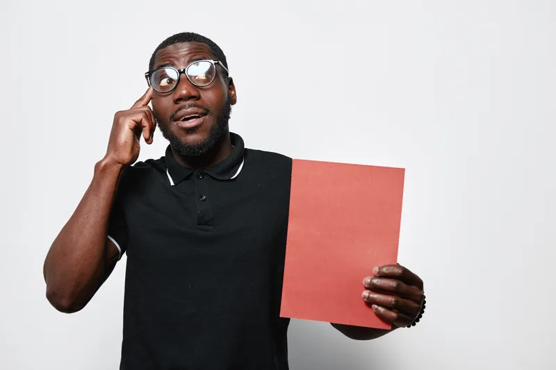 African American man with dark skin and glasses holding red paper while talking on smartphone, wearing black polo shirt, isolated on white background with thoughtful expression.