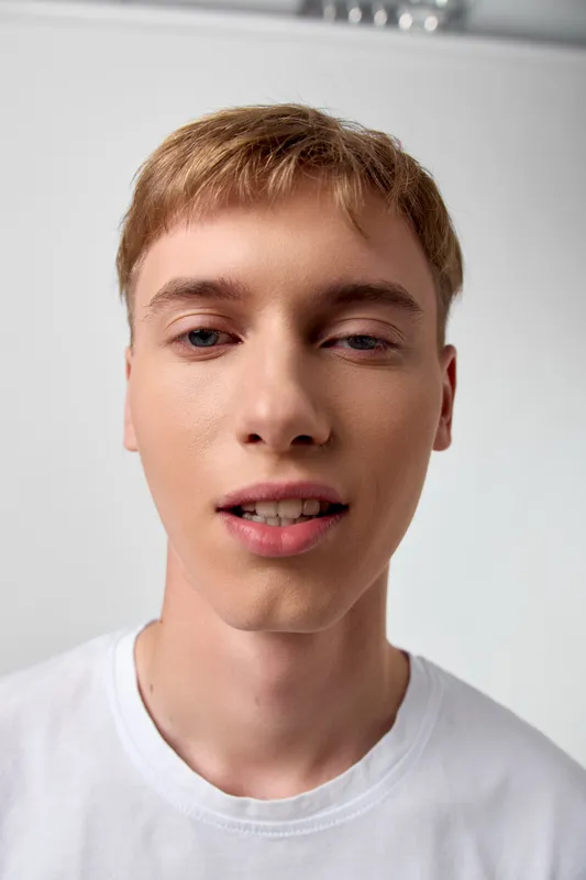 Close up of a young man in a white t-shirt, smiling softly indoors, showcasing natural light and clean skin tone for casual lifestyle and selfie style