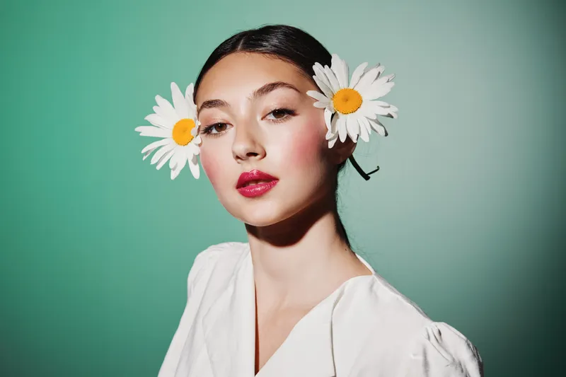 Young woman with daisies in her hair looks confidently into the camera. Clean makeup, glossy red lips. Minimalist fashion portrait on green background. Beauty and style concept.