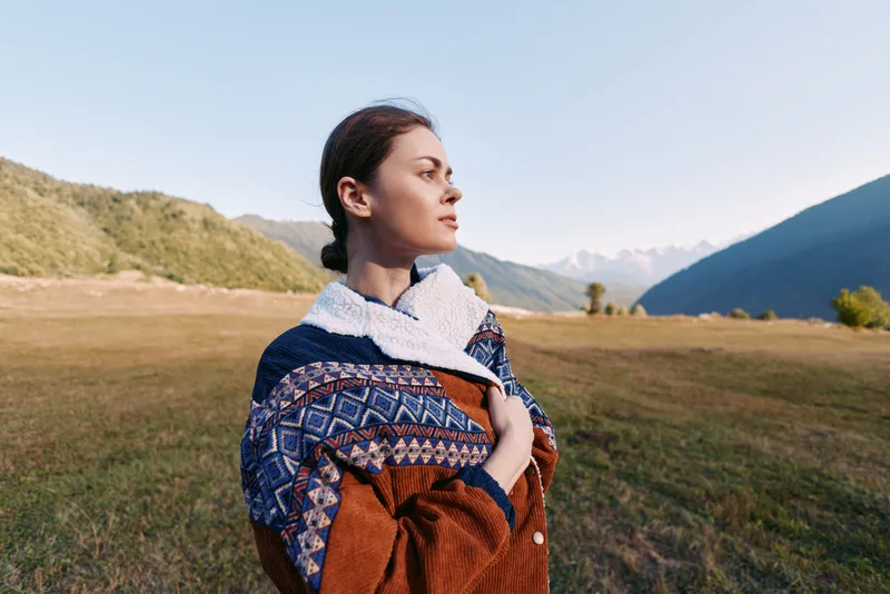 Woman portrait in mountains field wearing patterned scarf and warm sweater, side profile gaze toward horizon in nature meadow, peaceful autumn outdoor scene with soft sunlight.