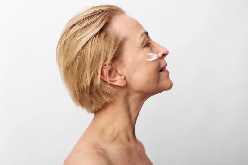 Mature woman with short blonde hair applies white face cream on cheek, smiling gently. Clean skin, skincare routine, self-care concept on white background with natural light.