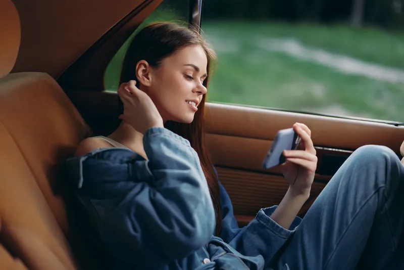 Young woman enjoying her smartphone in a car, wearing casual denim clothing, smiling and relaxed with a natural green background.