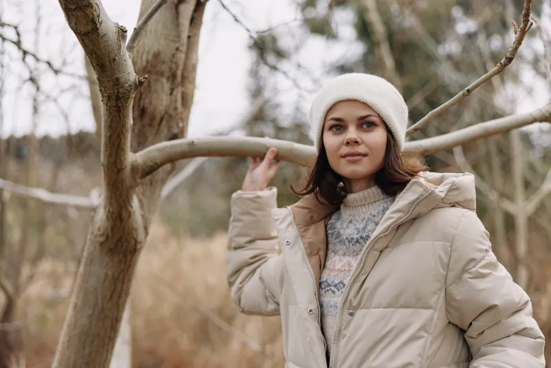 Woman in a winter coat standing outdoors among leafless trees, wearing a knit sweater and beanie, calm portrait in cold weather, natural light, soft tones, tranquil outdoor scene for fashion and