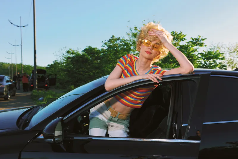 Woman leaning out of a sleek car, striped colorful top, retro film color mood, outdoor sunny scene, casual fashion pose, stylish snapshot with confident expression.