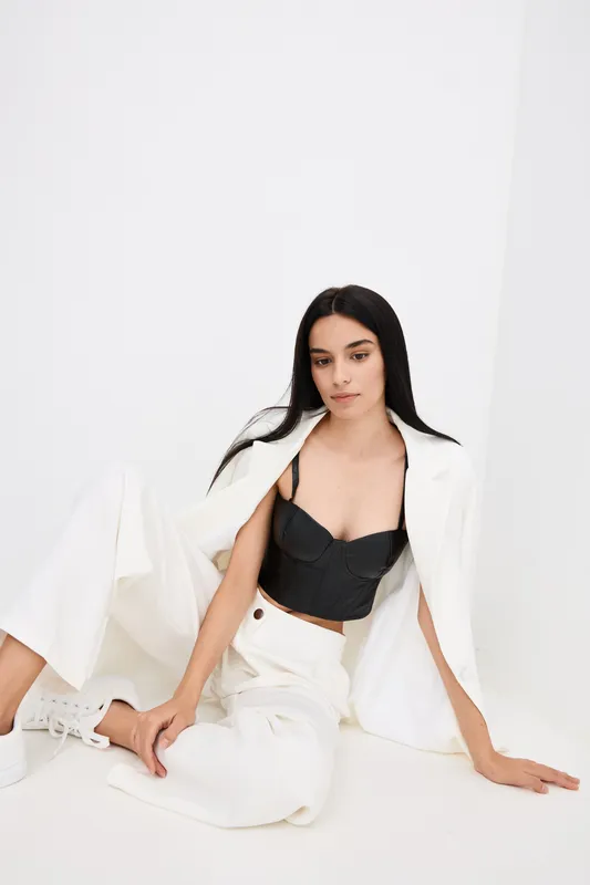 Young woman wearing stylish black bustier and white suit jacket with wide pants sits relaxed on floor against plain white background, showcasing modern fashion.