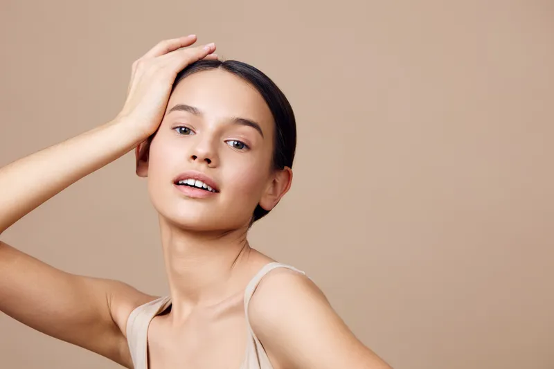 Portrait of a young, cheerful woman with brown skin and natural makeup, posing in a beige outfit against a warm, neutral background, ideal for beauty and skincare concepts