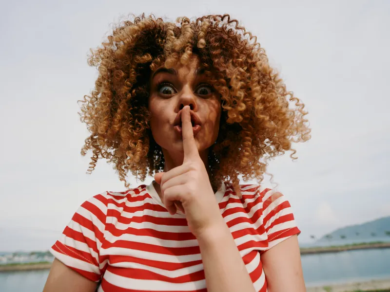 Close up portrait of a young woman with curly hair, making a shushing gesture, wearing a red and white striped shirt, outdoors with a bright sky and distant shoreline in the background.