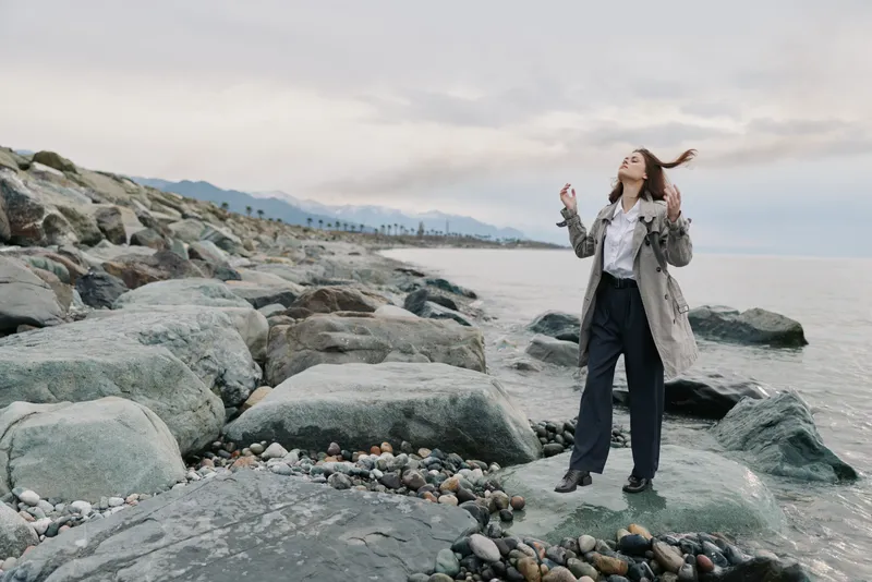 Young woman standing on rocky beach near calm sea with cloudy sky, wearing coat and trousers. Concept of nature, freedom, and outdoor lifestyle with peaceful atmosphere.