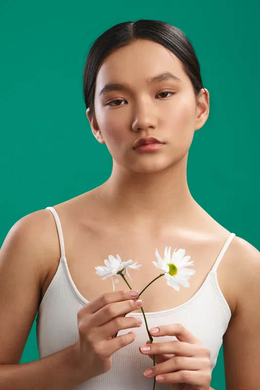 Asian woman stands before a vibrant green backdrop, gently holding daisies with a serene, thoughtful expression, creating a calm, natural portrait with soft lighting and minimal styling.