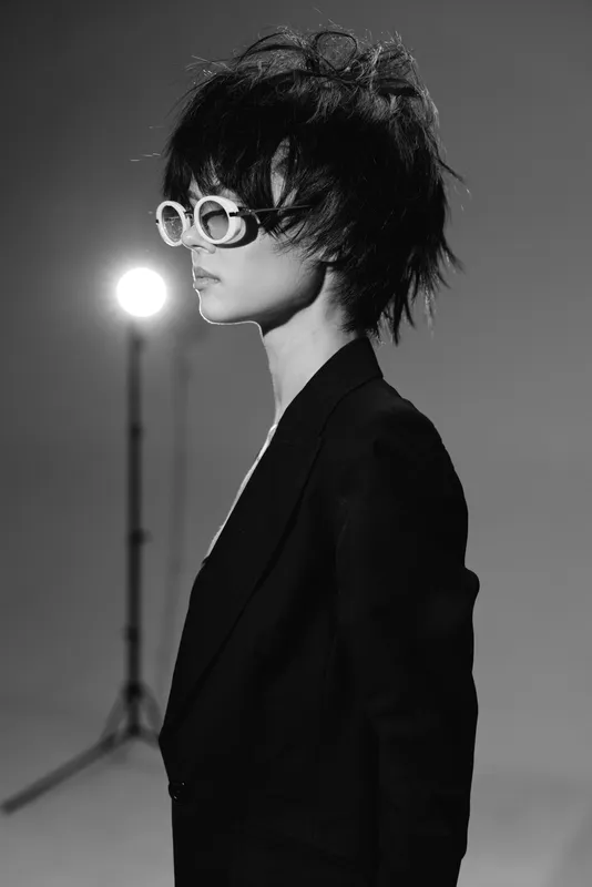 Black and white portrait of a stylish woman with sunglasses and short hair in dark clothing in studio lighting.