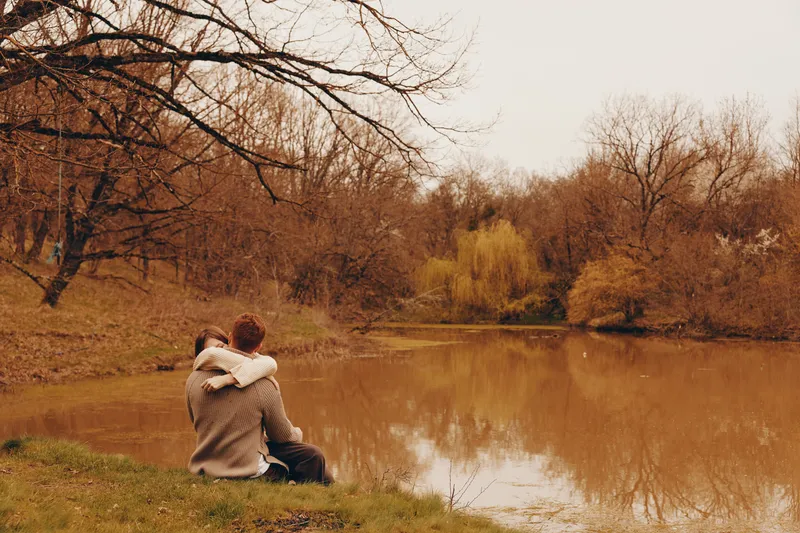 Couple sitting by a calm lake in autumn, embracing and enjoying the peaceful natural environment with bare trees and soft brown tones around them.