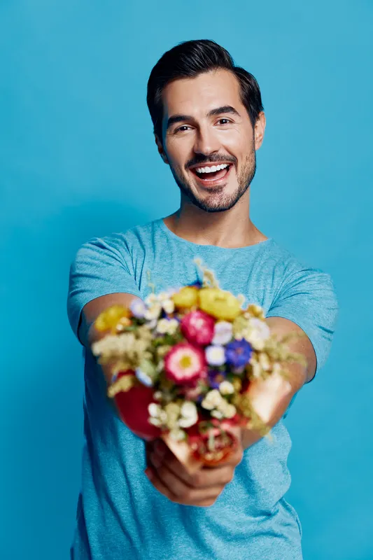 Cheerful smiling man holds vibrant bouquet of flowers while standing against bright blue background. Expressive portrait capturing joy and celebration.