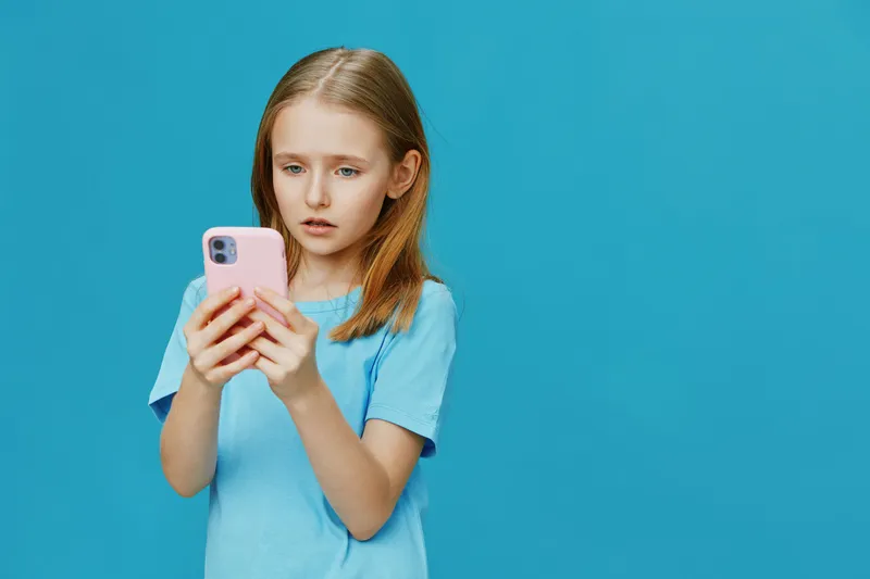Young girl looking surprised at her smartphone against a bright blue background, wearing a blue t shirt, expressing curiosity and engagement with technology