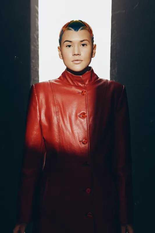 Young woman with short colorful hair wearing red leather coat standing between light and shadow, minimalist style, modern fashion, intense expression, urban background.