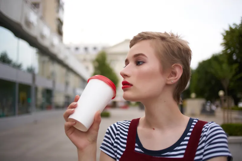 Женщина в очках на улице разговаривает по телефону, держа в руке стакан с напитком
