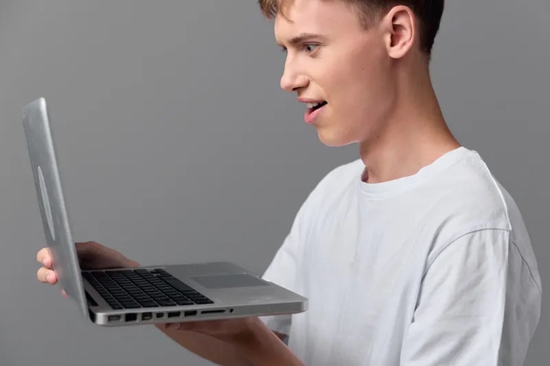 Man in a white tee holds a laptop and smiles at the screen, showcasing curiosity and modern tech use, casual work vibe, learning, remote tasks and digital creativity