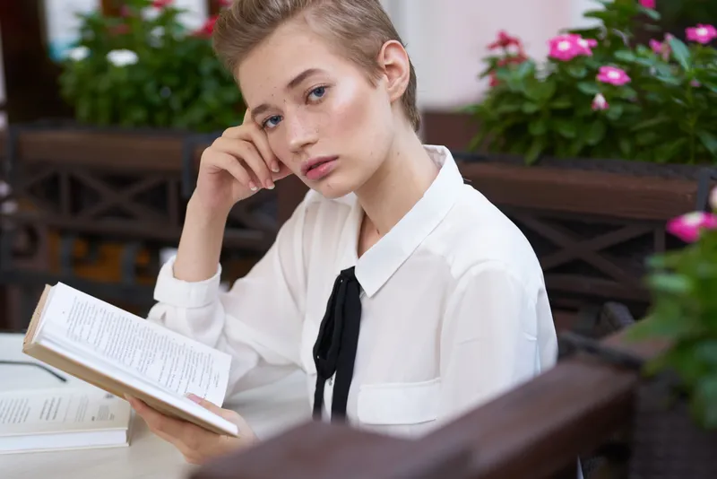 Умная женщина с книгой сидит за столиком в кафе, рядом розовые цветы, весенний макияж