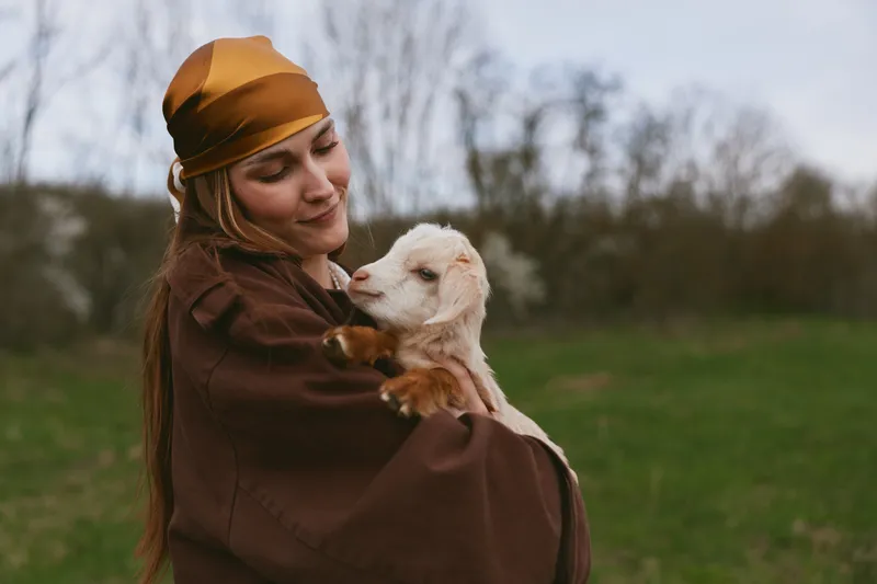 Woman in a brown headscarf cradles a goat kid in a grassy field, showing tender bonding outdoors. Rustic countryside vibe, natural light, calm mood, wholesome lifestyle moment.