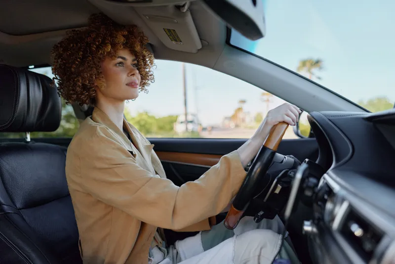 Calm woman driving a car, sunlit interior, curly hair, focused expression, casual jacket, window light, candid lifestyle moment, retro color tones, natural vibe