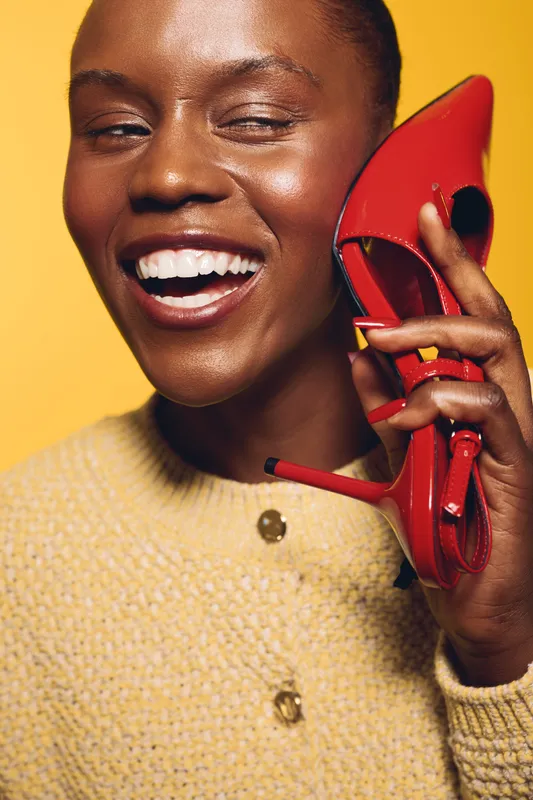 Smiling black woman with dark skin holds red high heel shoe near face on yellow background. Concept for fashion, beauty, style, and joyful emotions in studio portrait photography.