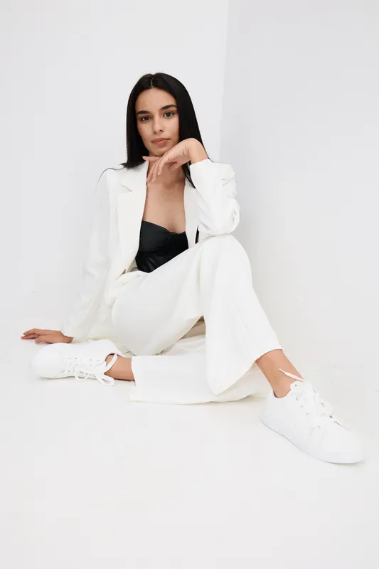 Young woman sitting on the floor wearing a white suit and sneakers, displaying a relaxed and stylish pose against a plain white background.