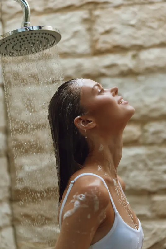 woman showering outdoors with closed eyes feeling relaxed under running water, natural stone background, wet hair, peaceful moment in warm light
