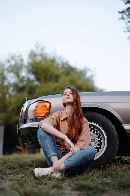 Stylish woman with long hair sitting next to a vintage car outdoors in casual attire, conveying a relaxed and thoughtful mood.