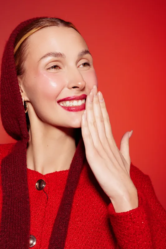Smiling woman with red lips and glossy skin wears red sweater and hood, covering part of her face with hand. Bright red background creates vibrant and joyful mood for beauty and fashion concept.