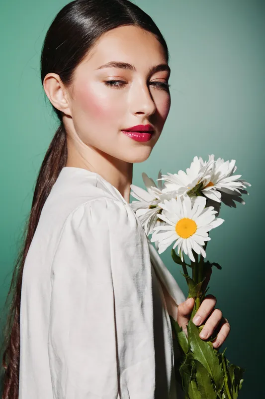 Young woman with sleek hair and vibrant pink lipstick holds white daisies, posing in high fashion studio light. Concept of beauty, elegance, and creative fashion portrait in soft green.