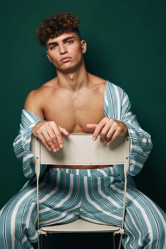 Young man with curly hair, shirtless wearing striped pants and unbuttoned shirt sitting backwards on chair against dark green background in studio portrait photo.