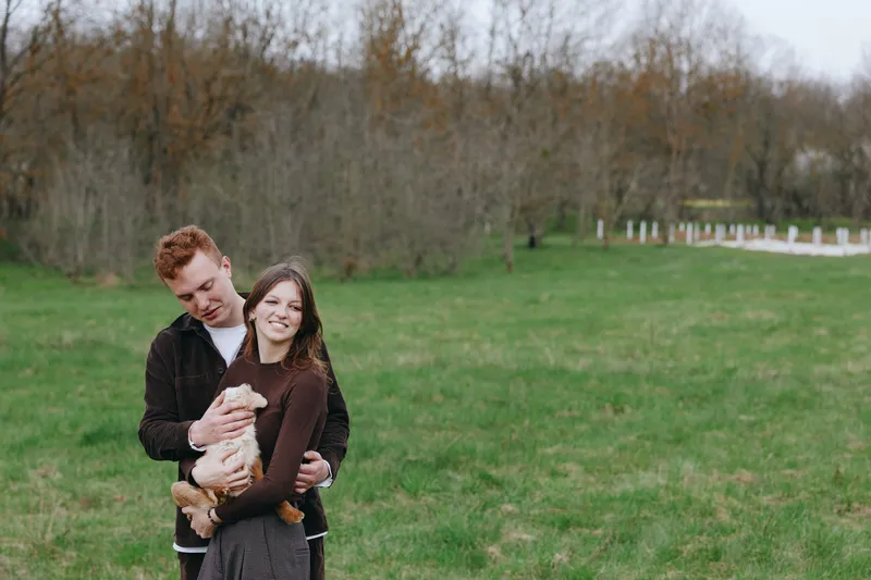 A young couple embraces outdoors as the woman holds a small baby goat, enjoying a peaceful moment in a green field with bare trees in the background during early spring.