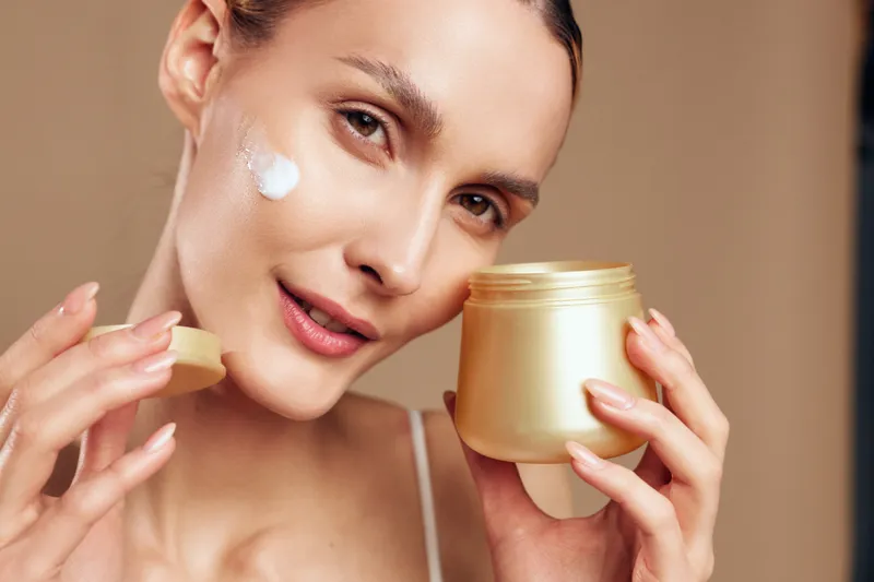 A young woman applies cream to her cheek while holding a jar of skincare product. The close-up shows natural makeup and smooth skin on a beige background.