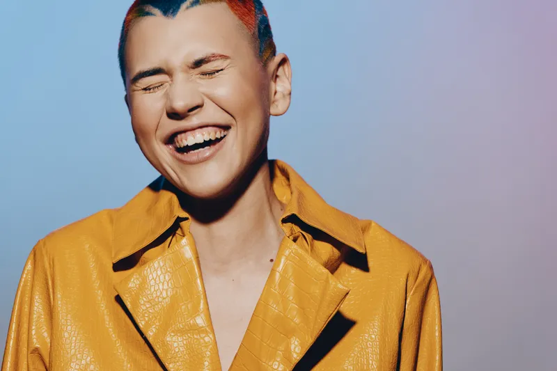 laughing woman with short hair wearing a bright yellow jacket, expressive joyful emotion, colorful gradient background, close-up portrait with vibrant lighting