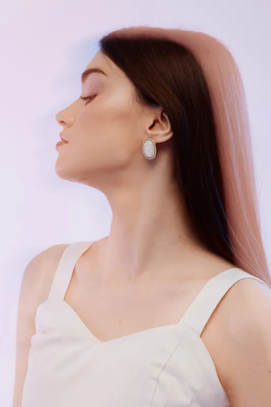 Profile of a young woman with long brown hair wearing a white dress and elegant earrings. Soft pastel background highlights beauty and fashion concept with natural makeup.