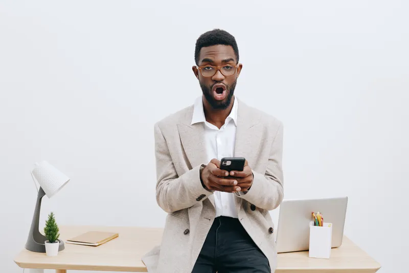 Smiling man using smartphone in office setting, surprised expression, casual beige blazer and white shirt, modern workspace with desk, laptop, stationery, minimal decor, clean background, business lif