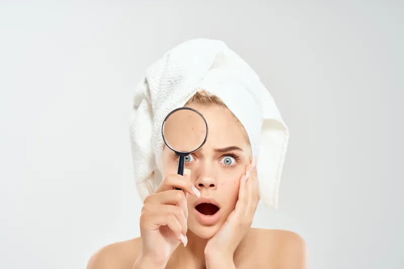 Крупный план лица симпатичной женщины с полотенцем на голове, дерматологическая лупа рядом с лицом