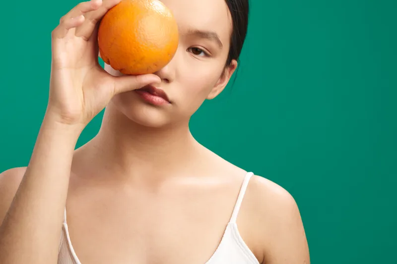 asian woman holds orange near eye against a green backdrop, calm expression, studio portrait highlighting fruit and curiosity, vibrant colors and a playful, contemporary mood.