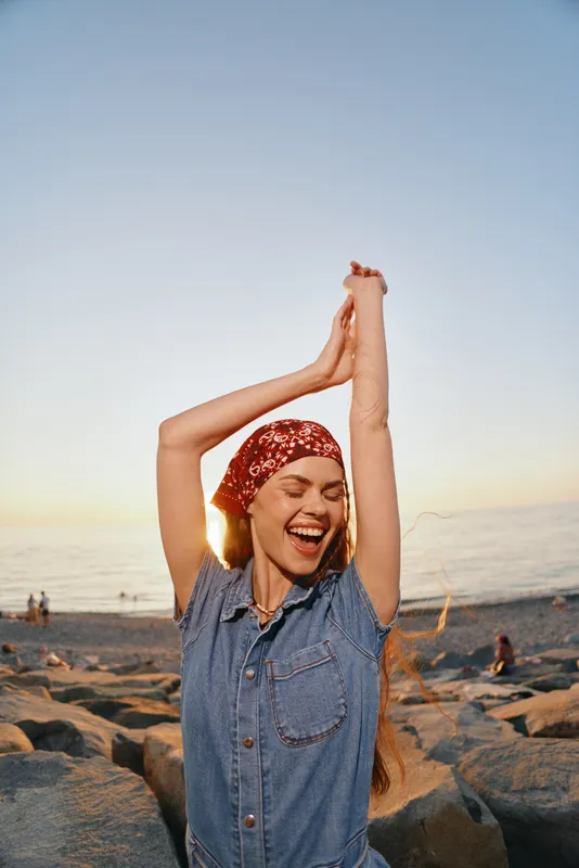 happy woman in modern boho-western denim street style smiling with raised arms during sunset on beach, film color lifestyle portrait showing joy and freedom