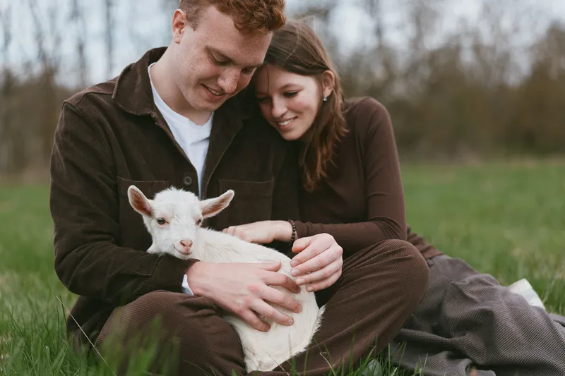 A couple sits on the grass outdoors, gently cuddling a white goat kid, capturing a rustic lifestyle moment and tender affection in a calm natural setting for a peaceful scene.